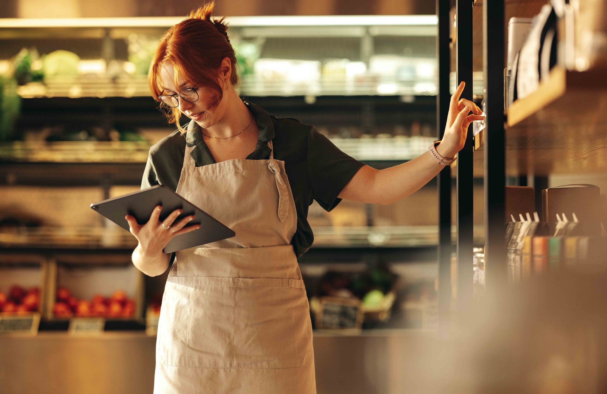 Woman working in a grocery shop wearing an apron and looking at a tablet device while reaching a shelf