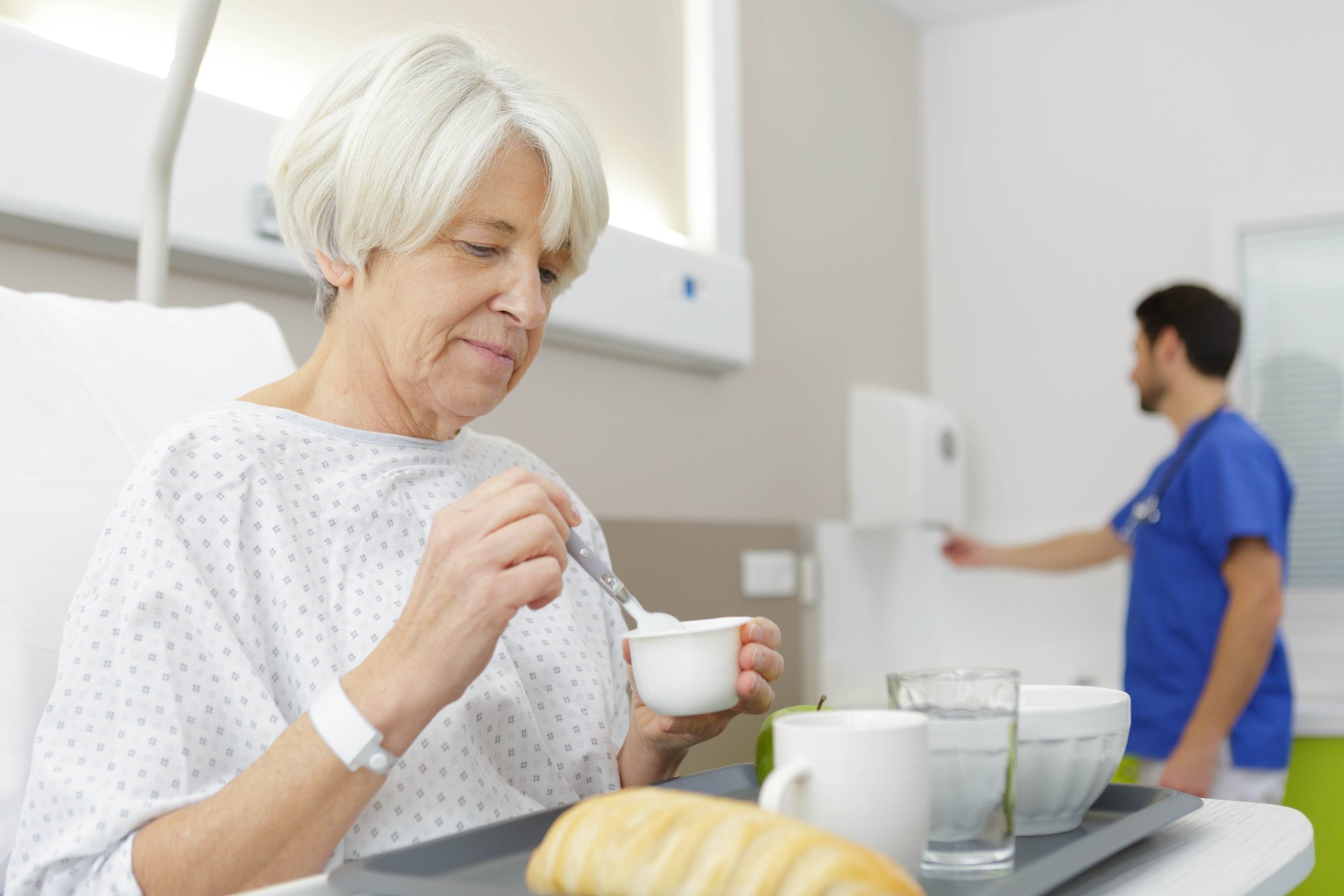 Woman in hospital bed holding a bowl and spoon in hands and nurse in the background