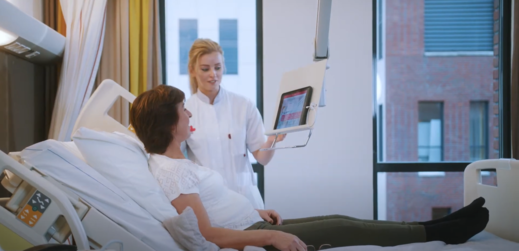 A nurse in a white uniform stands by a hospital bed showing a tablet to a patient lying in the bed. A professional medical facility setting.